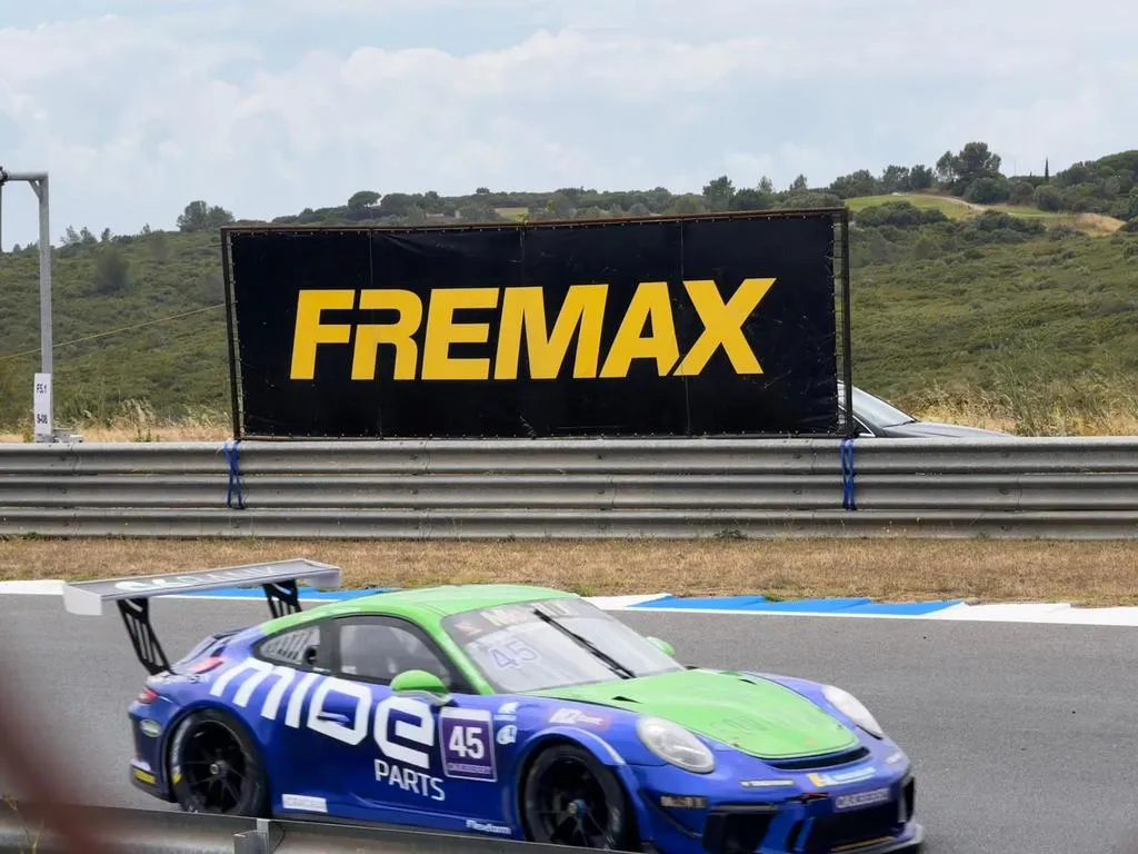 A imagem mostra um carro na competição da Porsche Cup Brasil e um outdoor da Fremax ao fundo. 
